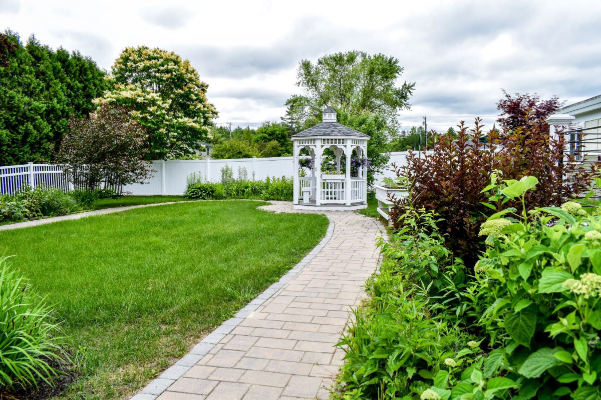 A beautiful garden with a brick pathway leading to a white gazebo surrounded by lush greenery.