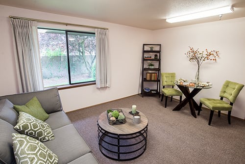 A cozy living room with modern furnishings, featuring a sofa, coffee table, bookshelf, and window view.