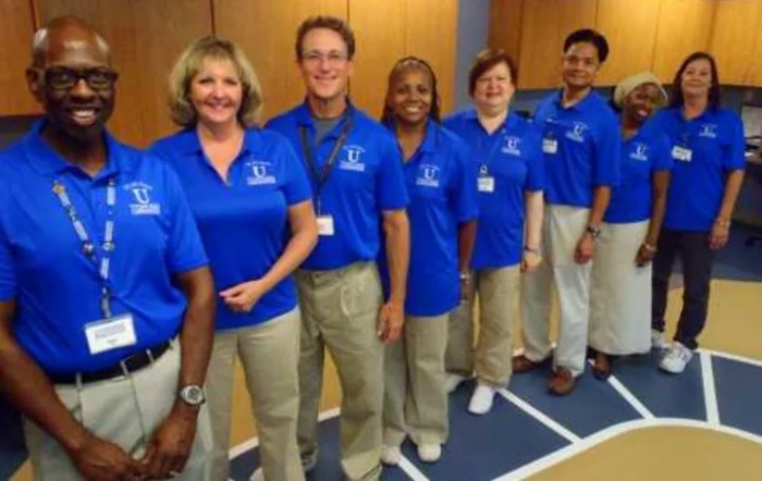 A group of six people wearing matching blue shirts and khaki pants, standing in a line and smiling.