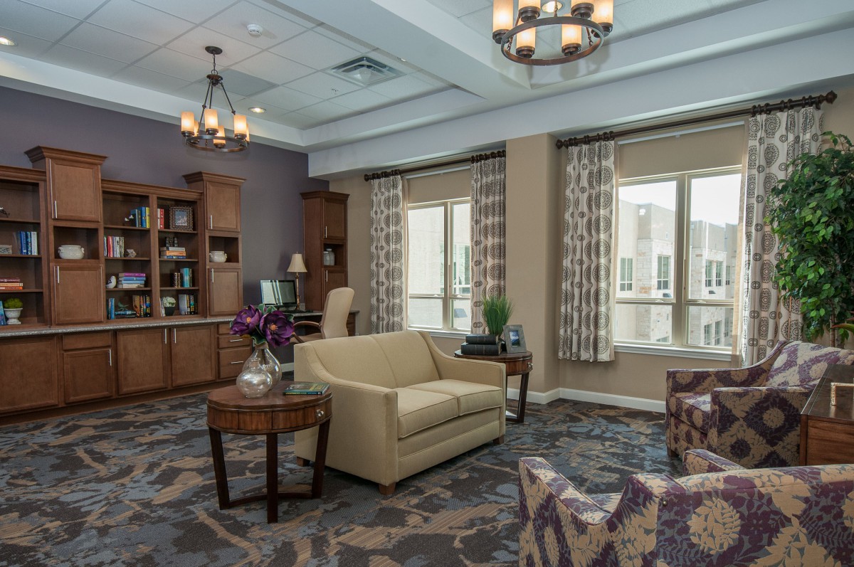 A cozy living room with elegant furniture and decor, featuring a sofa, armchair, coffee table, bookshelves, and large windows with curtains.