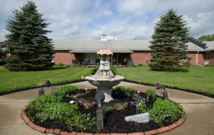 A garden with a central fountain surrounded by flowers and greenery, in front of a brick building.