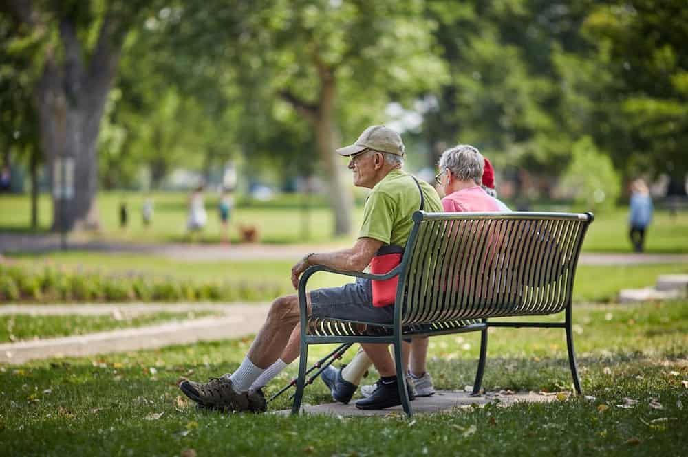 An elderly couple sits on a park bench, enjoying a sunny day outdoors.