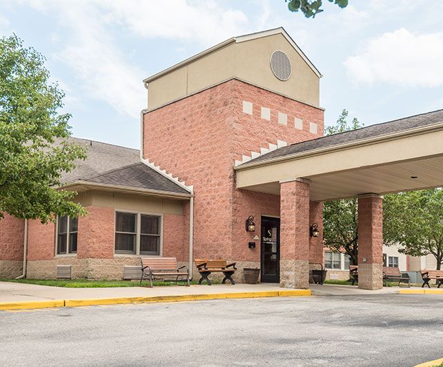 A brick building with a covered entrance and benches outside, surrounded by trees.