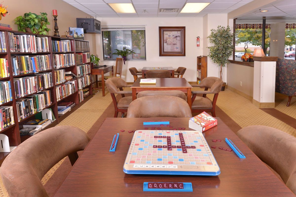 A cozy library room featuring a Scrabble game set up on a table surrounded by chairs, with shelves of books lining the walls.