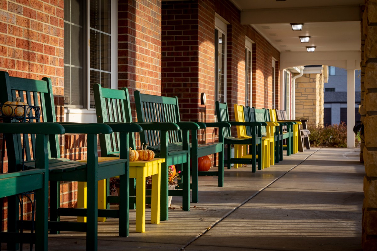 A row of colorful benches and chairs with pumpkins outside a brick building.