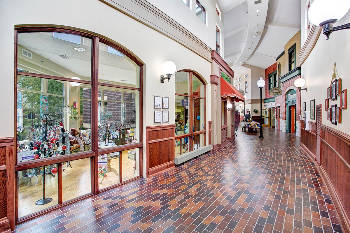 A spacious indoor corridor of a shopping center with brick flooring and large windows showcasing various shops.