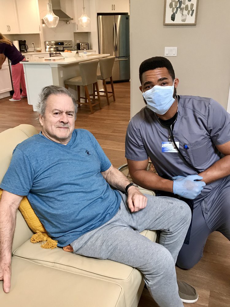 A healthcare worker wearing a mask and gloves is sitting beside an elderly man in a home setting.