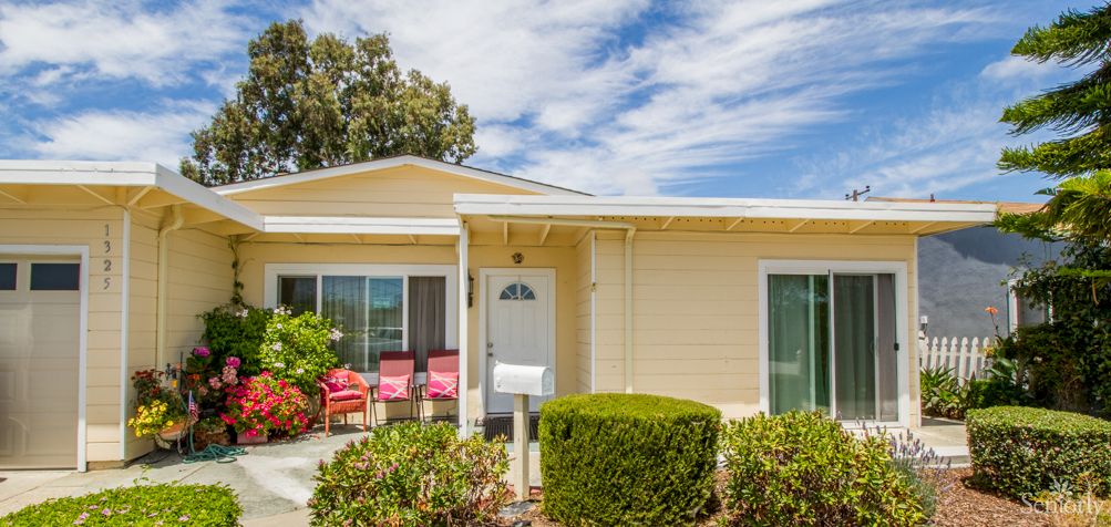 A cozy yellow house with a small front porch featuring red chairs, surrounded by neatly trimmed bushes under a partly cloudy sky.