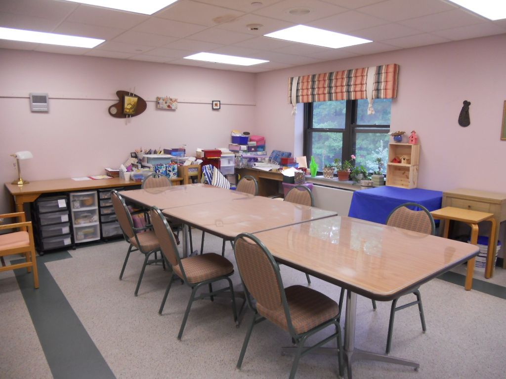 A well-organized classroom with tables and chairs arranged for activities, featuring craft supplies and natural light from the windows.