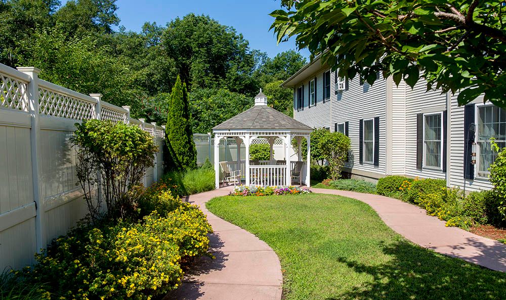 A well-maintained garden featuring a gazebo, surrounded by lush greenery and a paved pathway leading to a house.