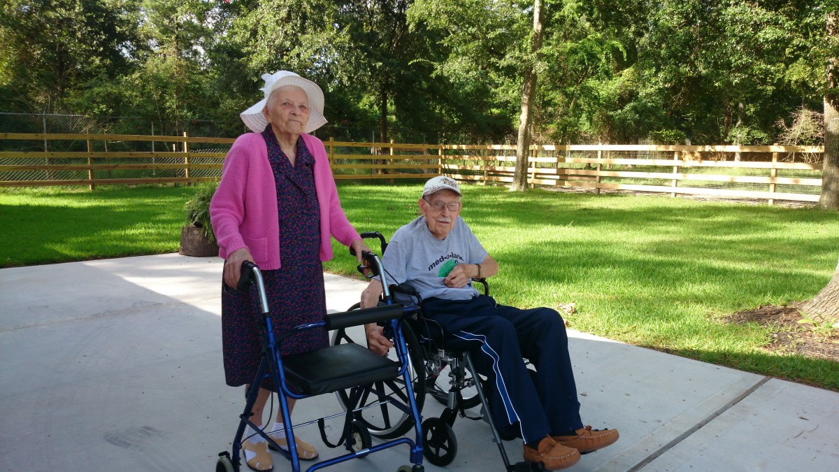 An elderly couple enjoying a sunny day outdoors, with one using a walker and the other in a wheelchair.