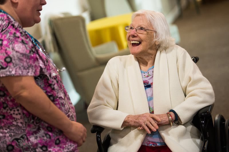 An elderly woman in a wheelchair is laughing and conversing with a caregiver indoors.