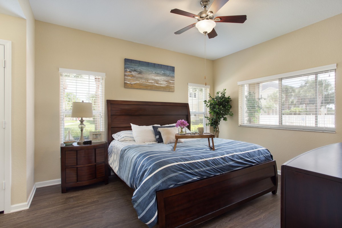 A cozy bedroom with a large bed, adorned with a blue striped comforter, flanked by wooden bedside tables. Sunlight pours in through several windows, and a ceiling fan hangs above.