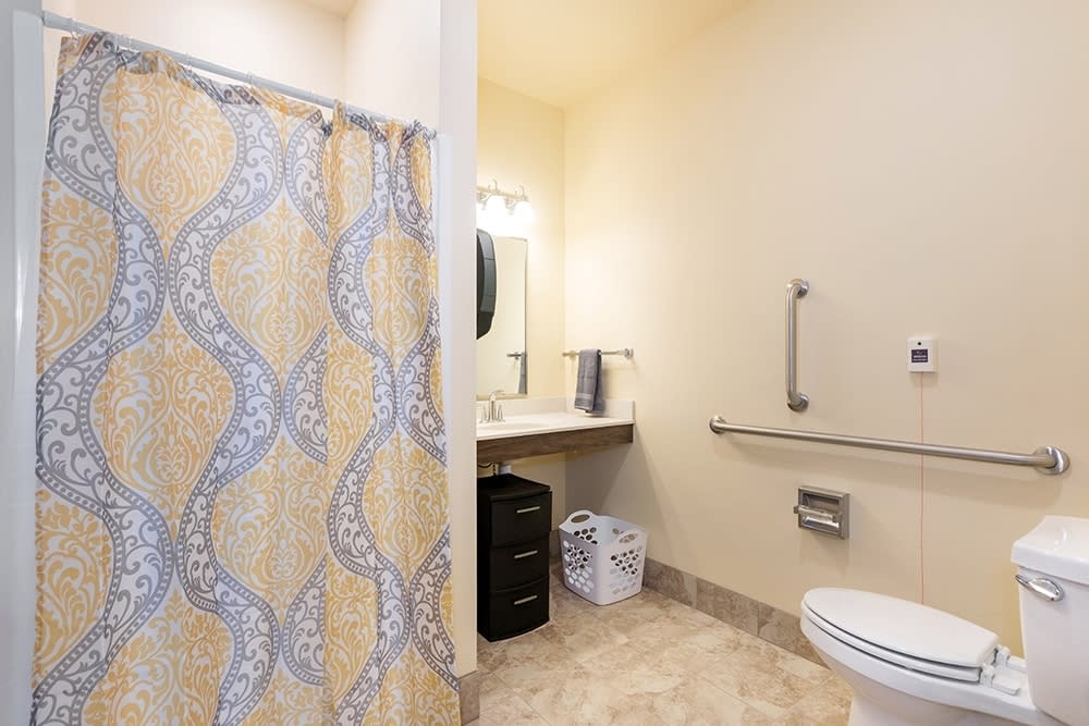 A clean and accessible bathroom featuring a shower curtain, sink, mirror, toilet, and grab bar.