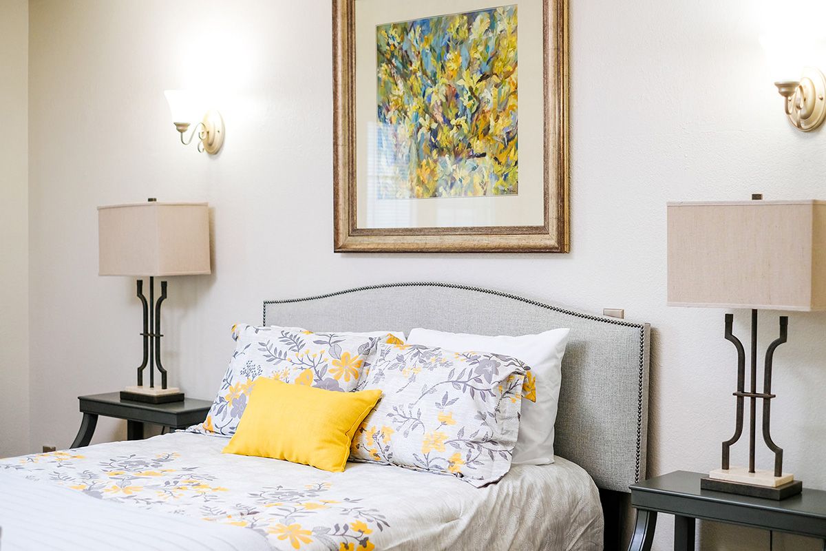 A well-decorated bedroom featuring a bed with floral bedding, a yellow accent pillow, stylish lamps, and framed artwork.