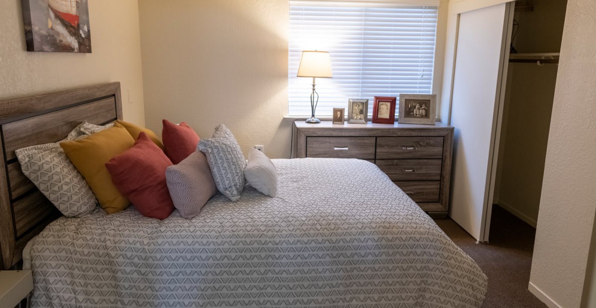 A cozy bedroom with a neatly made bed, soft pillows, and a lamp on the dresser by the window.