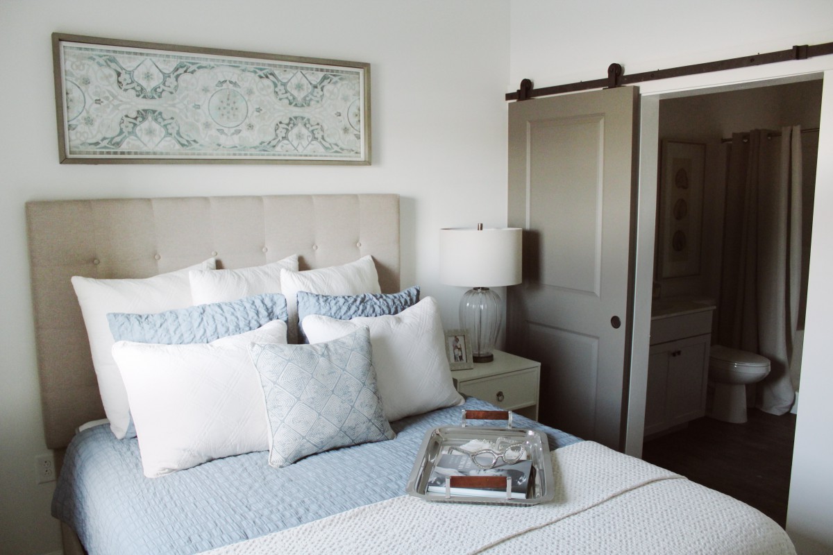 A minimalist bedroom with a neatly made bed, featuring blue and white pillows and a decorative wall piece. A lamp sits on a side table, and a sliding door is visible in the background.