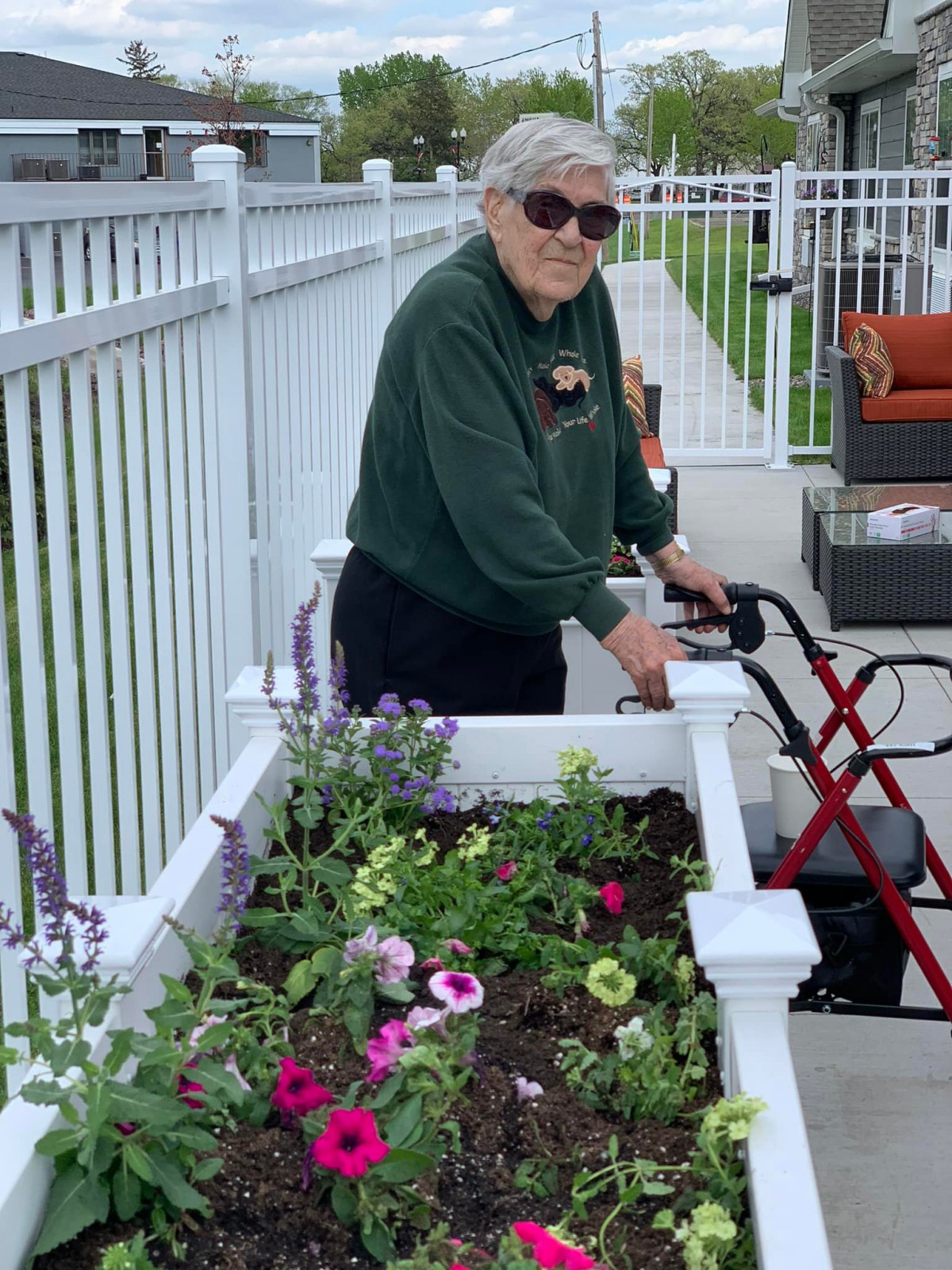 An elderly person using a walker is tending to a planter box filled with colorful flowers on a patio.