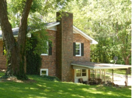 A brick house with a prominent chimney surrounded by trees and a lush green yard.