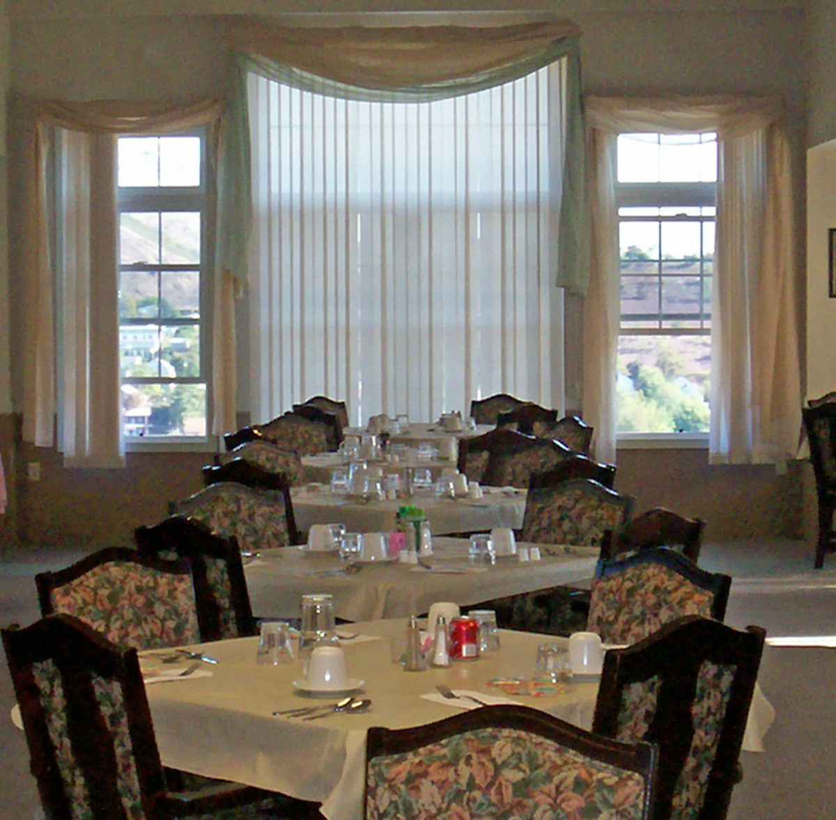 A dining room with elegantly set tables, featuring floral chairs and large windows framed by curtains.