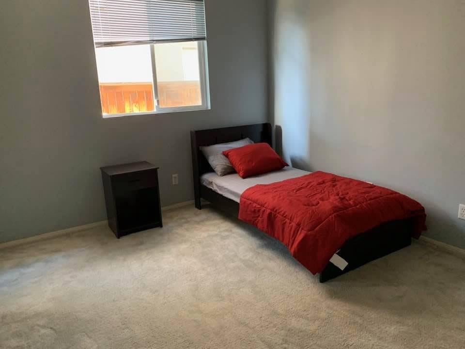 A minimalist bedroom featuring a single bed with red bedding, a nightstand, and a window providing natural light.