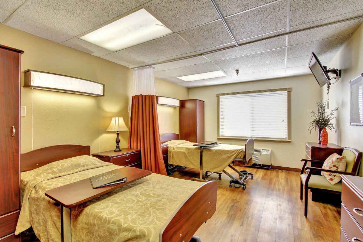 A well-lit hospital room with two beds separated by a privacy curtain and equipped with bedside tables and lamps.
