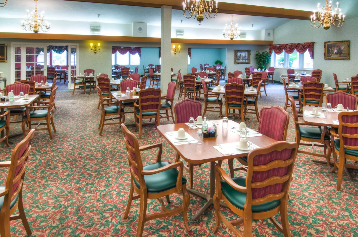A spacious dining area with elegantly set tables and chairs, featuring chandeliers and carpeted flooring.