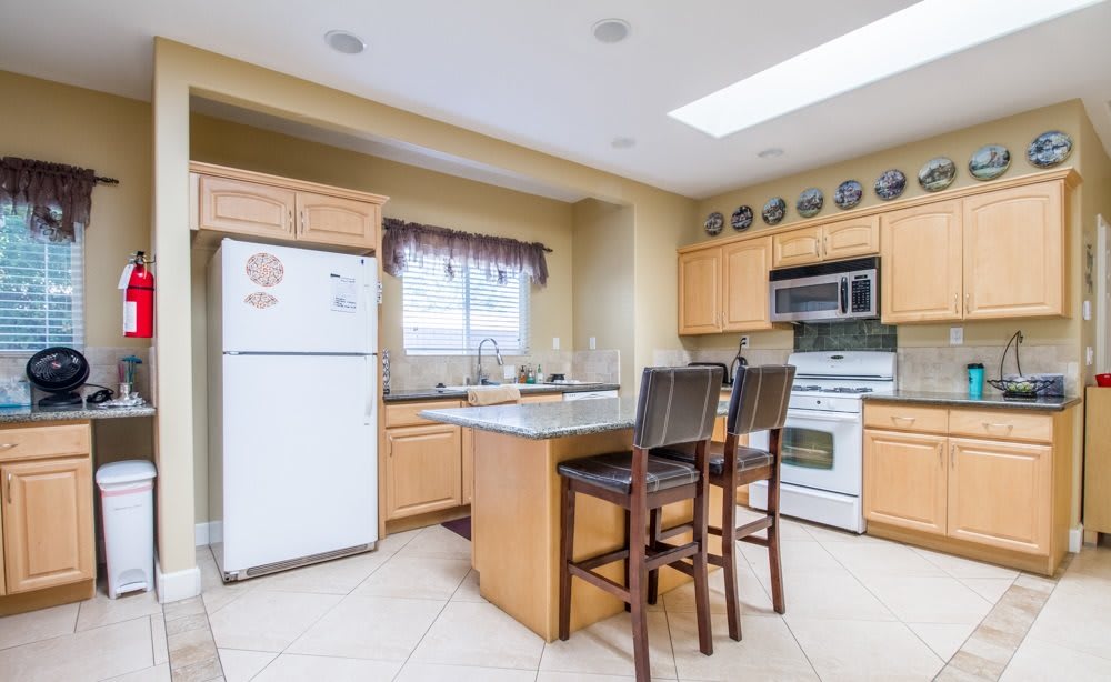 A modern kitchen interior with wooden cabinetry, featuring a fridge, stove, microwave, and a central island with bar stools.