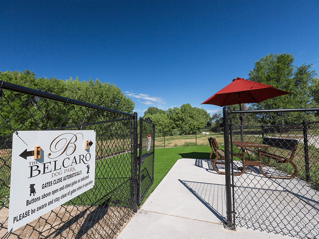 A fenced dog park with a red umbrella and seating area on a sunny day.