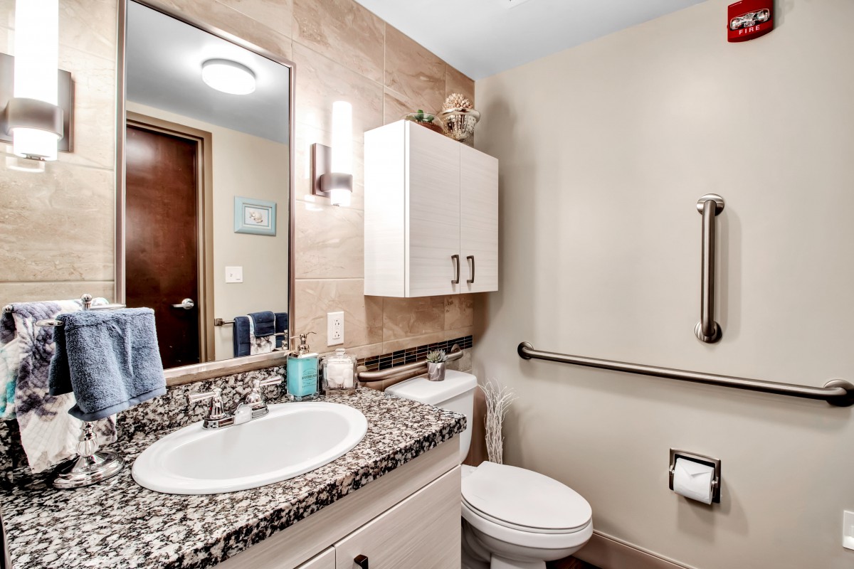 A modern bathroom featuring a granite countertop with a sink, a wall-mounted cabinet, and a toilet equipped with a handrail.