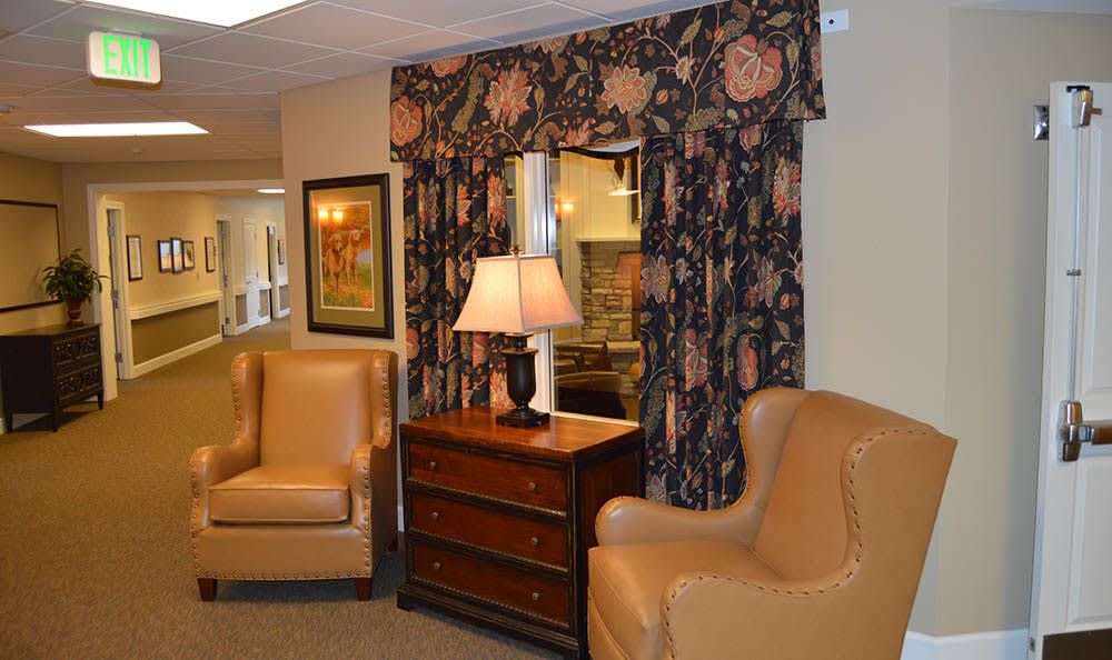 A cozy living room setup featuring a pair of beige armchairs, a wooden dresser with a lamp, and floral curtains.