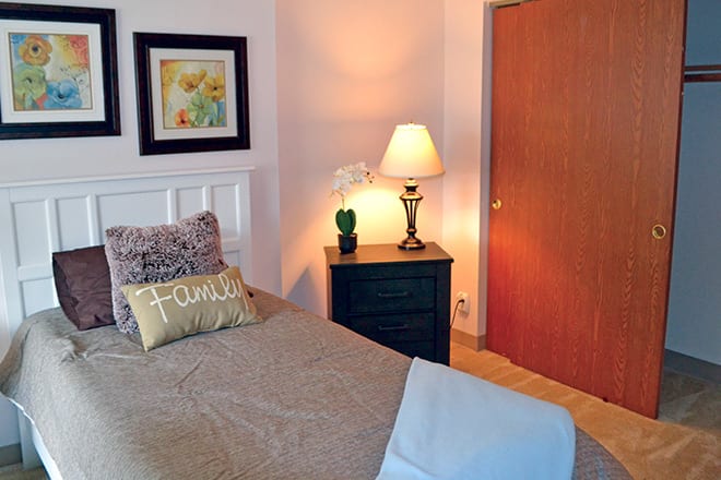 A cozy bedroom with a neatly made bed, featuring a decorative pillow, a nightstand with a lamp and plant, and framed artwork on the wall.