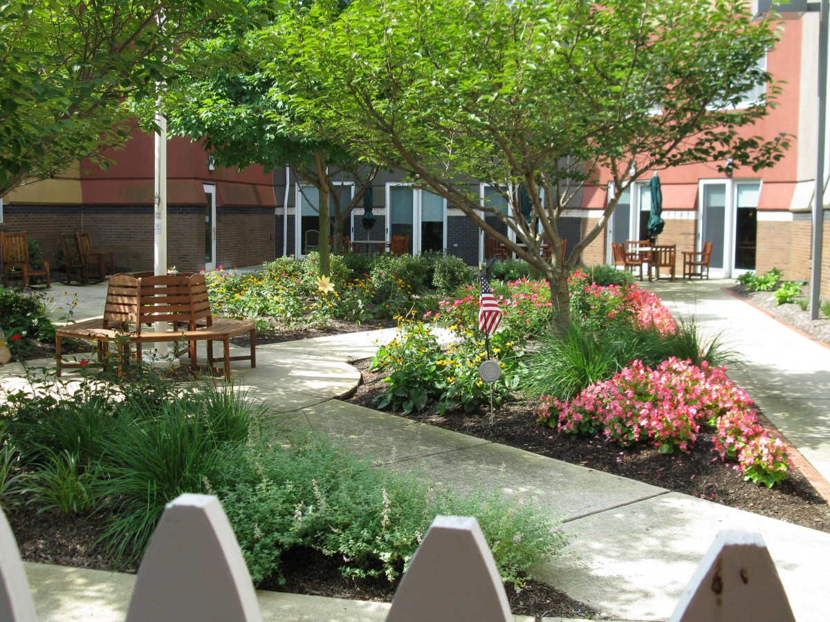 A peaceful garden with a pathway, surrounded by colorful flowers and trees, featuring wooden benches for relaxation.