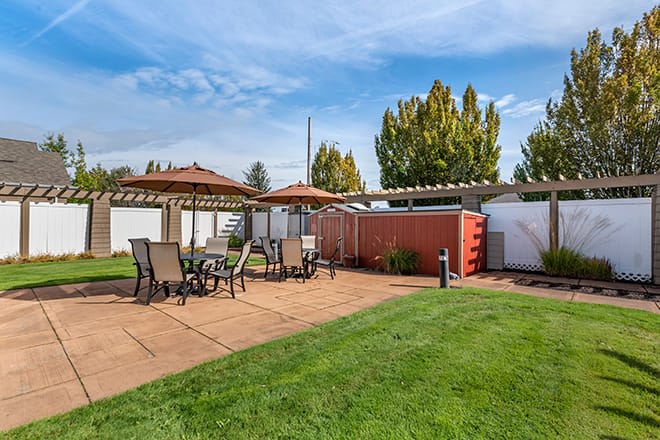A sunny outdoor patio area with tables, chairs, and umbrellas set against a well-maintained lawn.