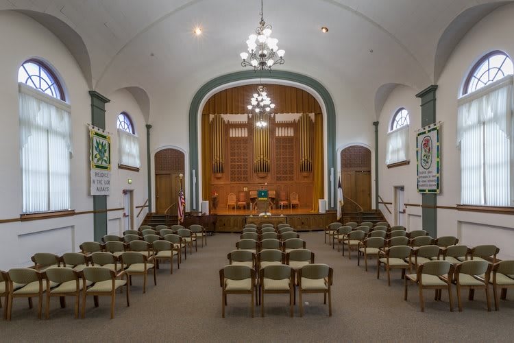 Interior view of a church with rows of chairs facing a raised stage area, featuring chandeliers and arched windows.