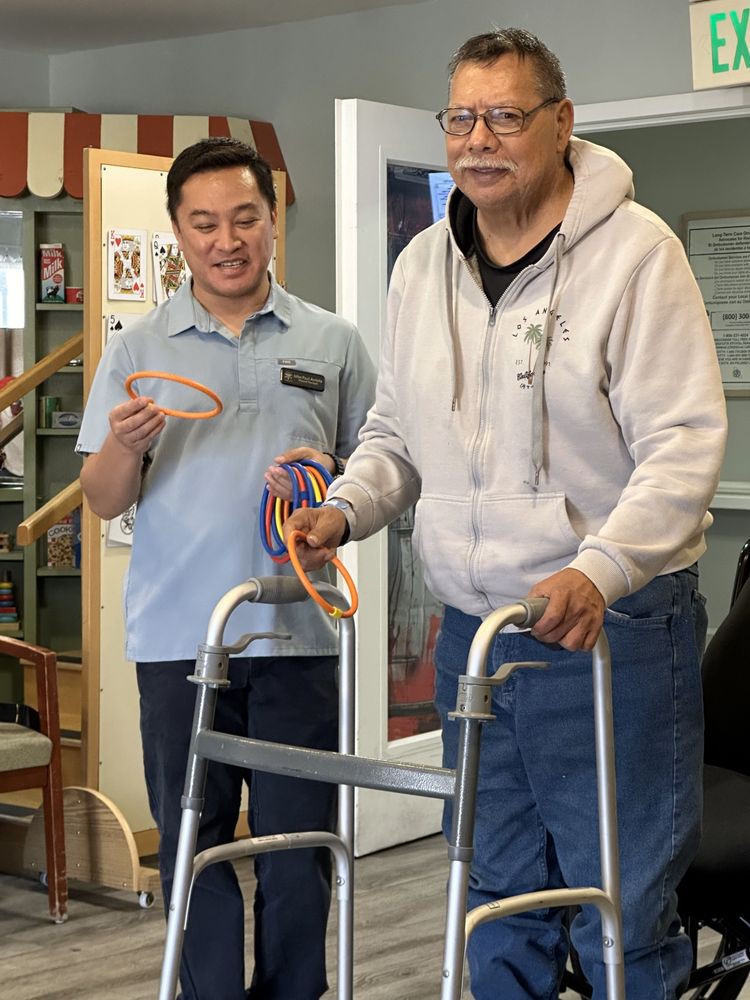 An elderly person with a walker receives assistance during a rehabilitation exercise session indoors.