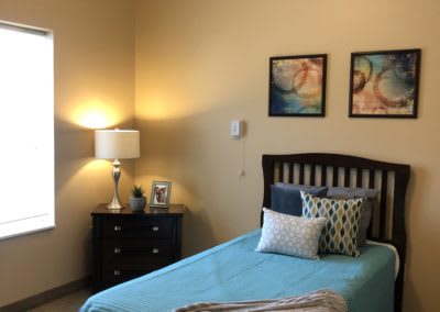A cozy bedroom with a single bed, decorated with colorful pillows and paintings, alongside a nightstand and lamp.