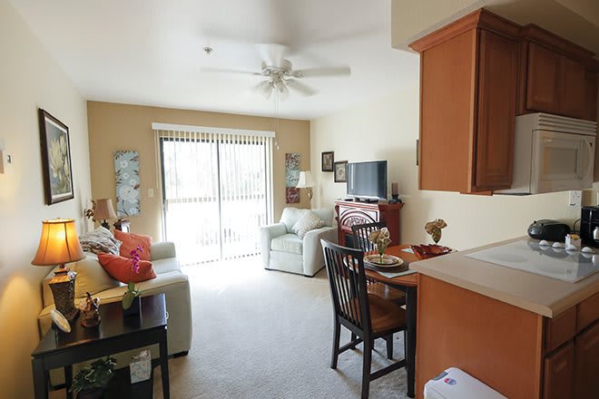 A cozy living room with a sofa, television, and dining area, featuring warm decor and natural light from large windows.