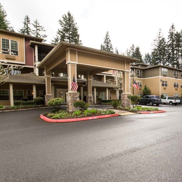 A view of a large building entrance with a driveway and parked cars, surrounded by trees under an overcast sky.