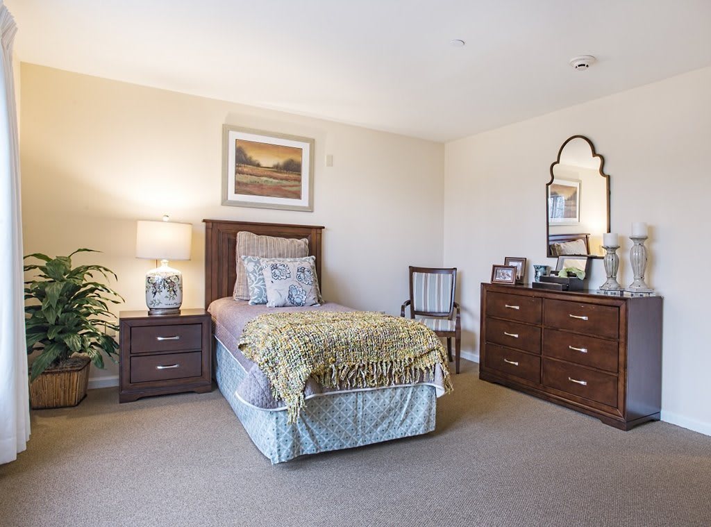 A cozy bedroom featuring a single bed, bedside table with a lamp, and a wooden dresser with a mirror.