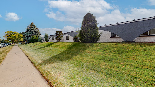 A peaceful landscape featuring well-maintained grass, trees, and modern buildings under a clear sky.