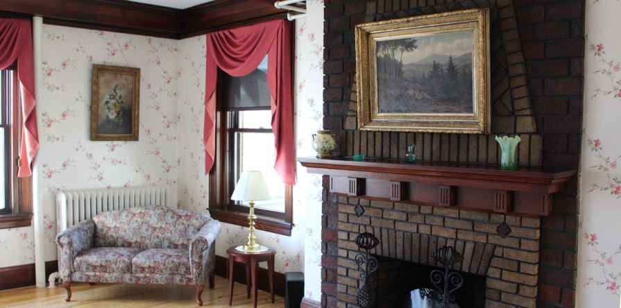 A cozy living room featuring a brick fireplace, adorned with a framed painting and red curtains.