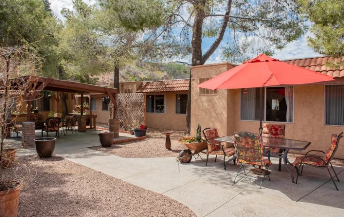 A serene courtyard patio with outdoor furniture and a red umbrella, surrounded by trees and an adobe-style house.