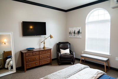 A cozy bedroom with a TV mounted on the wall, a wooden dresser, an armchair, and a large window.