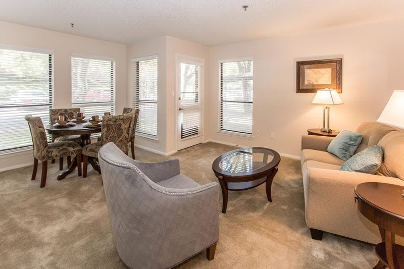 A cozy living room with a sofa and armchair, complemented by a dining area and ample natural light from multiple windows.