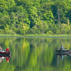 Two boats on a serene lake surrounded by lush green trees, with their reflections visible on the calm water.