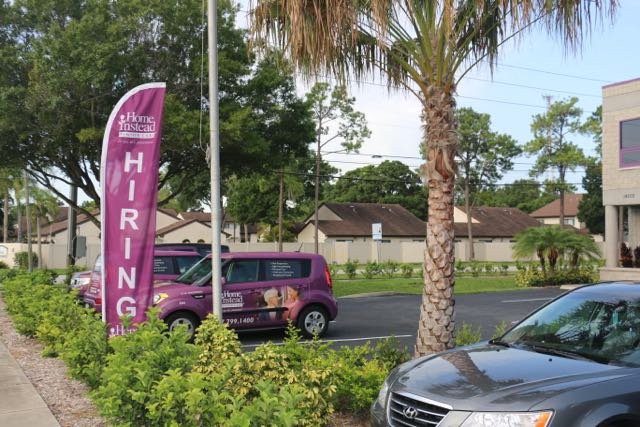 A 'Hiring' sign is placed outdoors near a road with a parked car and palm tree.