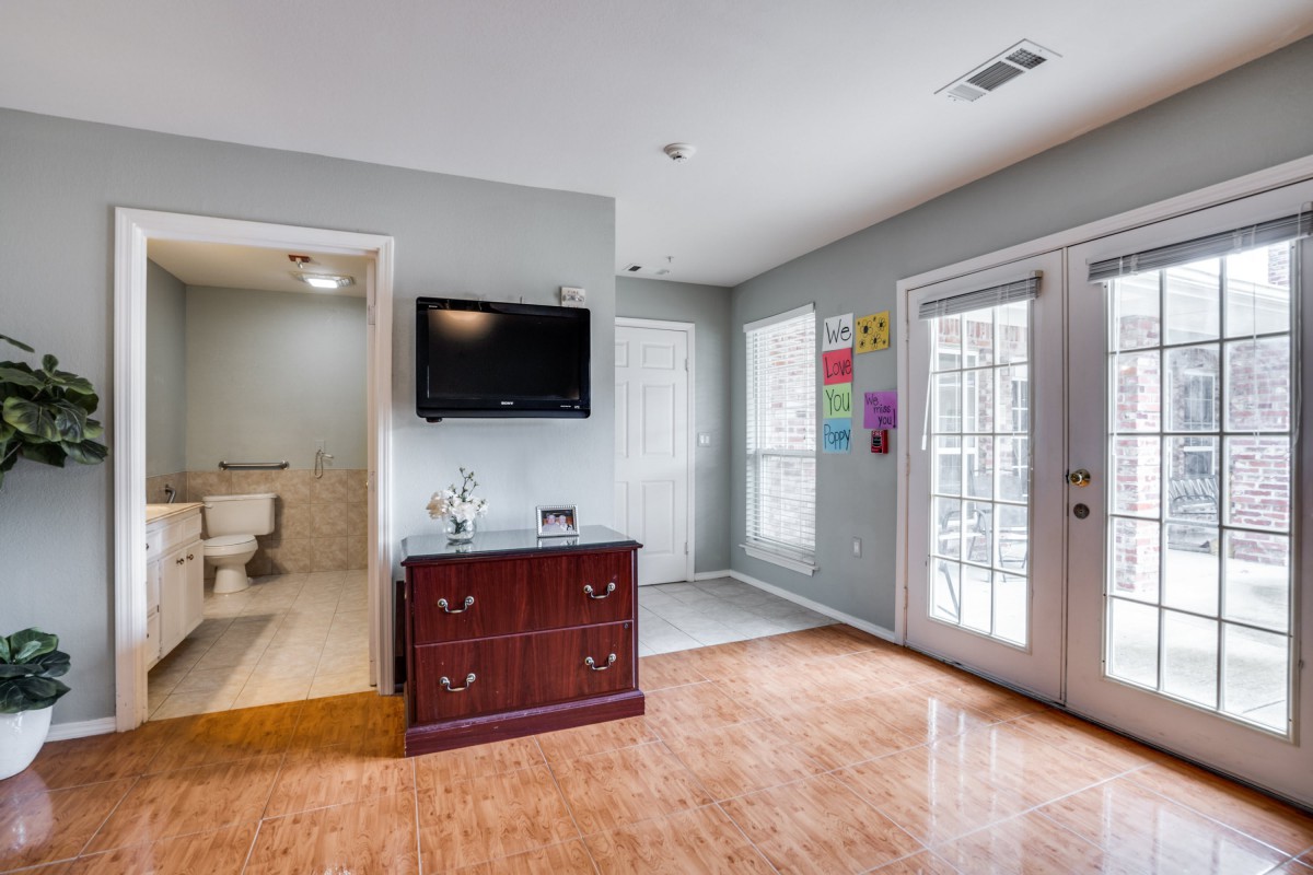 A cozy living area featuring a wall-mounted TV, wooden dresser, and bright artwork on the walls. The room connects to a bathroom and has a glass patio door letting in natural light.