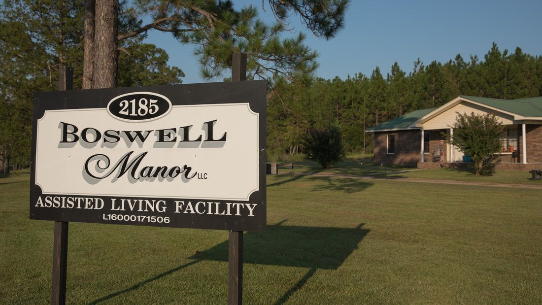 Sign for Maxwell Manor, a living facility, located in a grassy area with trees and a building in the background.