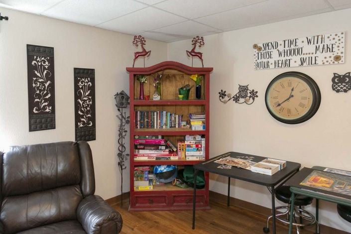 A cozy living room corner featuring a red bookshelf filled with books and games, alongside decorative wall art and a clock.